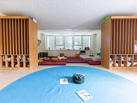 Ein Schlafraum mit einem großen, offenen Regal zur Aufbewahrung der Schlafausstattung; im Hintergrund liegen Matratzen mit Kissen und Kuscheltieren,