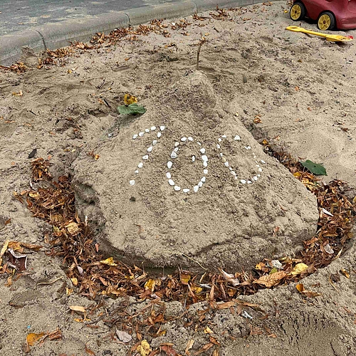 Ein Vulkan aus Sand mit der Zahl 100 in Steinen gelegt und einem Graben mit Blättern gefüllt um ihn herum