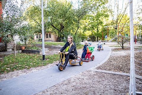 Eine pädagogische Fachkraft und Kinder befinden sich im Außenspielbereich; die Fachkraft fährt mit einem Dreirad voraus, fröhliche Kinder folgen ihr auf ihren eigenen Dreirädern. Im Hintergrund sind weitere Spielzeuge.