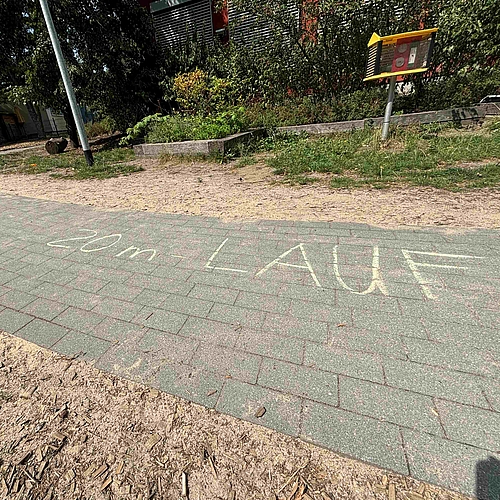 auf dem Bild ist ein grauer Steinweg im Garten des Kindergartens zu sehen. Auf dem Weg wurde mit gelber Kreide "20m-Lauf" geschrieben