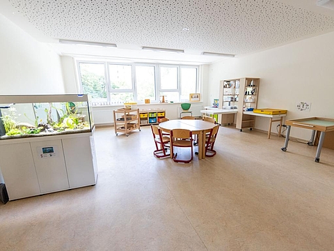 Ein Raum mit einem Aquarium auf der linken Seite, einem runden Tisch in der Mitte und einem Regal im hinteren Bereich, das mit Forschermaterialien wie Mikroskopen ausgestattet ist.