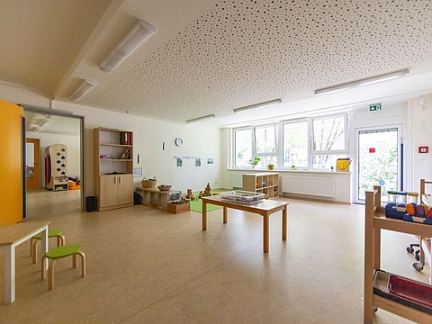 Ein großer Raum mit Tischen in der Mitte; im hinteren Bereich stehen Regale und es befindet sich eine kleine Bauecke für Kinder.
