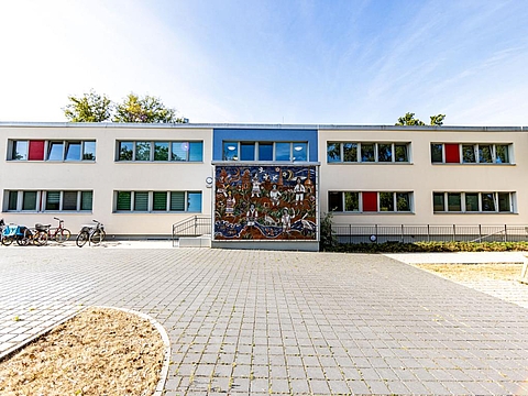 Außengelände des FRÖBEL-Kindergartens Sausewind mit einem Gebäude, das sich über das Gelände erstreckt; die Fassade wechselt zwischen blauen und beigen Farbbereichen, einige Fensterrahmen sind rot und blau, und der Eingangsbereich ist mit einer farbenfrohen Wandmalerei gestaltet.