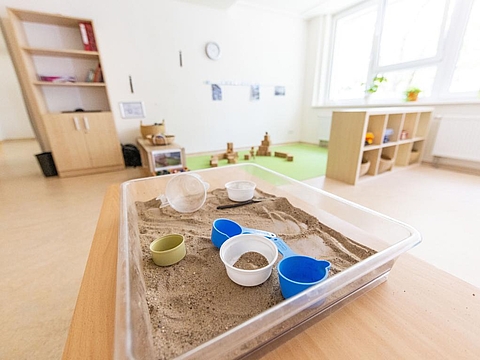 Ein großer Sandkasten im Raum, in dem viele Eimer verteilt sind; im Hintergrund stehen Regale und eine kleine Bauecke lädt zum kreativen Spielen ein.